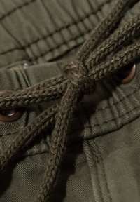 Tela verde oliva con textura visible, que presenta un cordón trenzado, costuras reforzadas y ojales de metal marrón para ajuste.