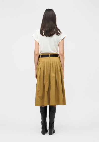 Γυναίκα με μακριά σκούρα μαλλιά, φοράει λευκό κοντομάνικο τοπ, μουσταρδί πλισέ φούστα μέχρι το μέσο της γάμπας, μαύρη ζώνη και μαύρες μπότες μέχρι το γόνατο, απεικονίζεται από πίσω.