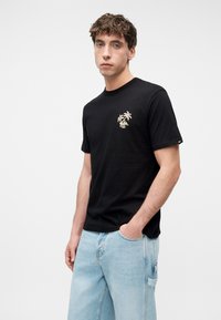 Νέος άνδρας που φοράει μαύρο μπλουζάκι με σχέδιο φοίνικα στο στήθος και ανοιχτό μπλε τζιν, στέκεται με το ένα χέρι στην τσέπη, μπροστά σε λευκό φόντο.