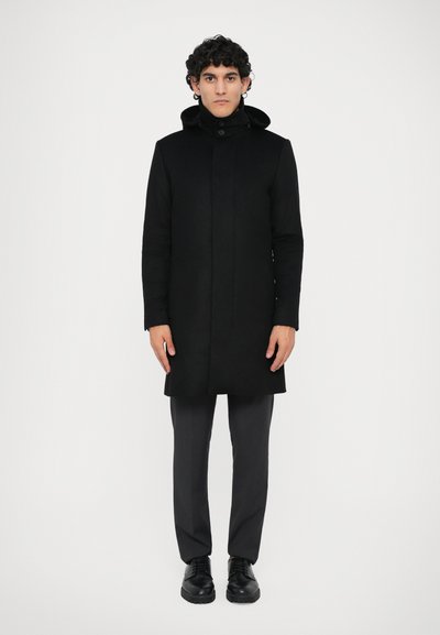 Zwarte jas met hoge kraag en afneembare capuchon; heeft een rechte snit, lange mouwen en een minimalistisch ontwerp. Zwarte broek en schoenen.