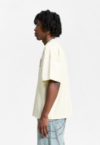Lysegul oversized t-skjorte med korte ermer, med brodert tekst. Båret med lyseblå jeans, prydet med kjedeakenter.