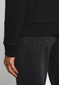 Felpa nera con polsini a coste, abbinata a jeans grigio scuro con tasca posteriore e trama in denim visibile.