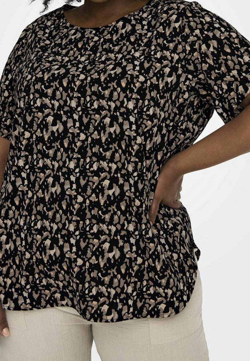 Femme portant un haut à manches courtes à motif abstrait noir et beige, avec un pantalon beige clair, main posée sur la hanche.