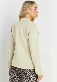 Beige denimjacka med krage, figurnära design och sidoknappar, bärs över svarta och bruna leopardmönstrade byxor. Strukturerad tyg synlig.