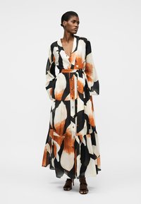 Vrouw die een lange, met knopen verzierde bloemrijke jurk draagt met zwarte, witte en oranje abstracte patronen, staand met handen in de zakken en zijdelings kijkend.