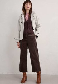 Světle šedá dešťová bunda přes vínový pletený svetr a pruhované tričko. Hnědé kordové kalhoty s širokými nohavicemi a hnědé kotníkové boty na podpatku.