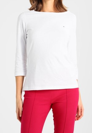 Frau trägt ein weißes Langarmshirt und eng anliegende, leuchtend rote Hose, steht vor einem einfarbigen hellen Hintergrund.