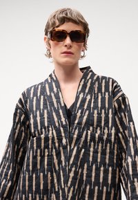 Jachetă neagră de tip kimono, cu un model abstract în nuanțe de bej și crem, având decolteu în V, mâneci largi și buzunare frontale. Modelul poartă ochelari de soare supradimensionați.