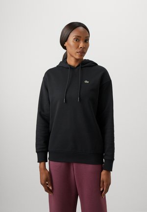 Svart hettegenser med snøring, ribbestrikkede mansjetter og kant. Har en liten grønn logo. Glatt tekstur på stoffet. Kombinert med burgunderbukser.