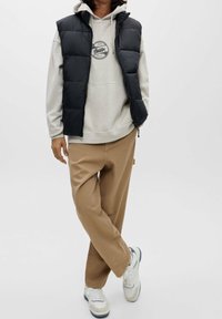 Svart vadderad väst över en ljusgrå huvtröja med logotyp, kombinerat med beige byxor och vita sneakers. Släta texturer; avslappnad design.