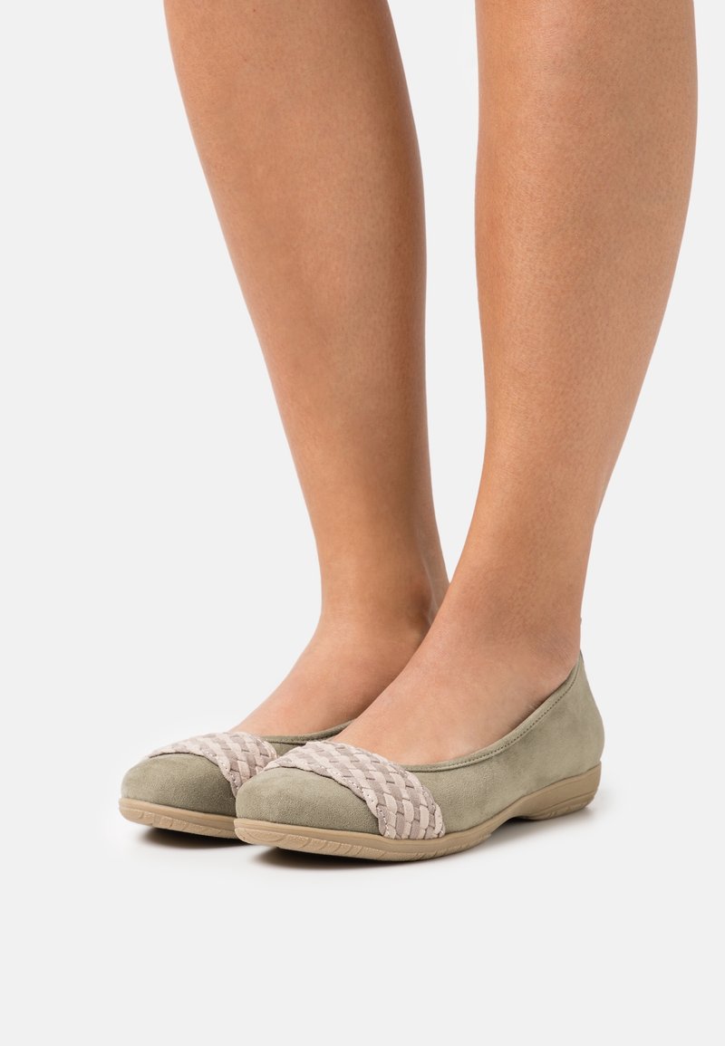 Jambes portant des chaussures plates vert olive clair avec un détail tissé beige sur le bout, debout sur un fond clair uni.