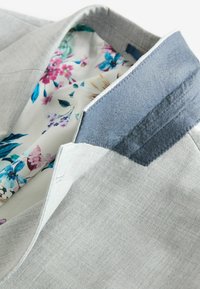 Blazer gris claro con un acabado texturizado. El interior presenta un forro con un patrón floral en rosa, azul y verde. El cuello tiene un detalle de fieltro azul.
