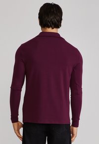 Bărbat purtând o cămașă strâmtă cu mâneci lungi, de culoare violet închis, și pantaloni negri, stând cu spatele spre un fundal simplu.