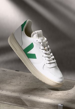 White sneaker made of leather, featuring a green rubber 'V' logo, perforated toe, and a textured cream rubber sole.