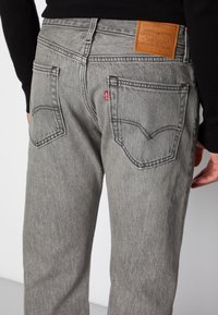 Harmaat farkut suoralla leikkauksella, joissa on kaksi taka-taskua, nahkainen tuotemerkkiylitys ja huomaamaton ompelutaito, käytetty lantiolla.