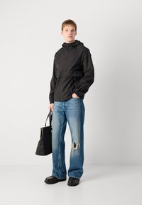 Zwarte hoodie-jack met een gestructureerd patroon, gecombineerd met versleten blauwe jeans en zwarte schoenen. Het model houdt een zwarte tas vast.