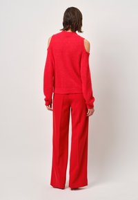 Rode, oversized trui met een off-shoulder ontwerp; zachte textuur. Gecombineerd met hooggewaisted, wijde rode broek. Minimalistische achtergrond.