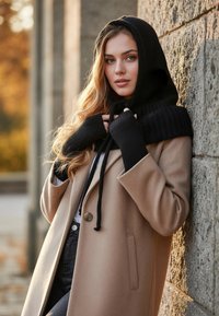 Cappotto in lana beige con un solo bottone, cappuccio nero a maglia con lacci e guanti neri senza dita. Muro di pietra e sfondo autunnale.