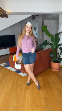 Jauna moteris, apsirengusi rožiniu megztiniu ir džinsine sijonu, stovi svetainėje, laikydama rankinę, šalia sofos ir didelio kambarinio augalo.