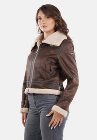 Chaqueta de cuero marrón con cuello y puños de piel de oveja, con cierre de cremallera y bolsillos laterales, terminada con una cintura ceñida con cinturón.