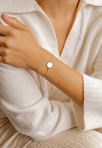 Bracciale d'oro con un pendente rotondo e liscio, caratterizzato da una delicata catena. La mano poggia su un morbido tessuto bianco dalla texture.