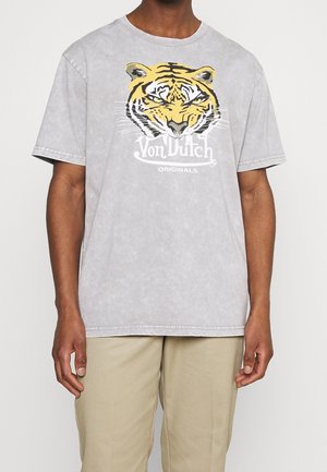 Homme portant un t-shirt gris clair avec un visage de tigre jaune et le texte « Von Dutch Originals » à l'avant, associé à un pantalon beige.