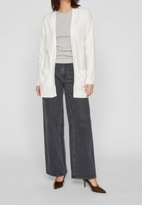 Cardigan bianco a maglia con chiusura aperta e due tasche frontali, indossato sopra una t-shirt grigia, abbinato a jeans larghi scuri e tacchi marroni.