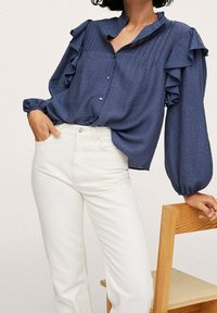 Navy button-up shirt with ruffled sleeves, textured fabric, and a high collar, paired with white jeans. Model poses with one hand in pocket.