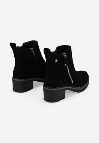 Par de botas estilo botín de ante negro con tacones bajos, cremalleras laterales y lengüetas, sobre un fondo blanco.