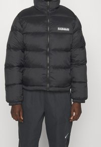Veste matelassée noire avec fermeture éclair à l'avant et logo Napapijri sur la poitrine, portée avec un pantalon Nike gris foncé.