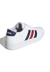 Bijela tenisica s tri horizontalne pruge u mornarsko plavoj i crvenoj boji. Napravljena od glatkog sintetičkog materijala s umetkom od pjene oko gležnja i dizajnom s vezanjem.