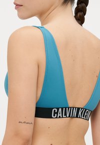Blå sport-bh med djup ryggdesign, som har en mjuk textur och ett kontrasterande svart elastiskt band med "CALVIN KLEIN" logotyp.