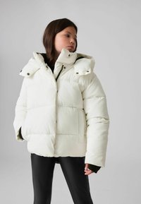 Vit pufferjacka med hög krage och dubbel huva. Har stora quiltade sektioner, svarta detaljer och sidofickor.