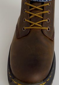 Bota de cuero marrón con cordones amarillos, punta redonda y costuras amarillas contrastantes a lo largo de la suela. Presenta ojales de metal y lados texturizados.