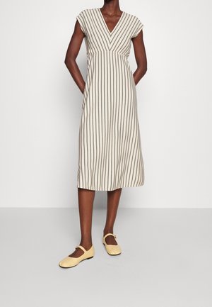 Femme portant une robe crème mi-longue avec des rayures verticales noires et des manches courtes, associée à des ballerines Mary Jane jaune pâle.