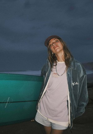 Kurtka sztruksowa w ciemnym odcieniu teal, haftowany t-shirt w jasnym lawendowym kolorze oraz szorty w paski. Akcesoria obejmują czapkę i kolczyki. W tle deska surfingowa.