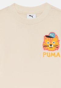 Camiseta de algodón beige con un colorido gato de dibujos animados con gorra y la palabra "PUMA" en letras naranjas y en negrita en el frente.