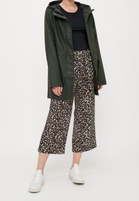 Person i en mörkgrön regnjacka, svart topp, beige leopardmönstrade vida byxor och vita sneakers, som står mot en enkel bakgrund.