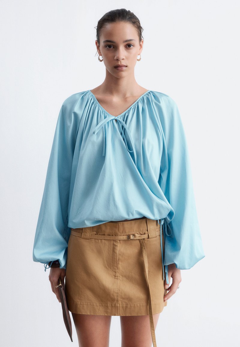 Femme aux cheveux attachés porte un chemisier bleu clair à manches longues et une jupe beige, tenant une petite pochette marron devant un fond clair.