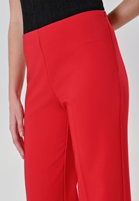 Pantaloni rossi a gamba larga realizzati in tessuto liscio, con vita alta, cerniera laterale e dettagli sottili sulla cucitura laterale.