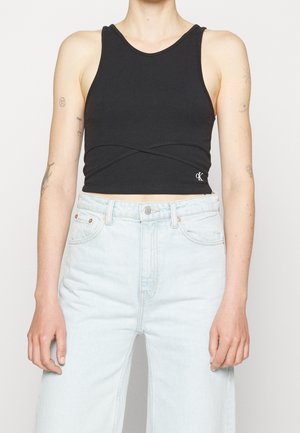 Černý krátký nátělník bez rukávů s překříženými švy, nošený s vysoce pasovanými světle modrými džínami na osobě.