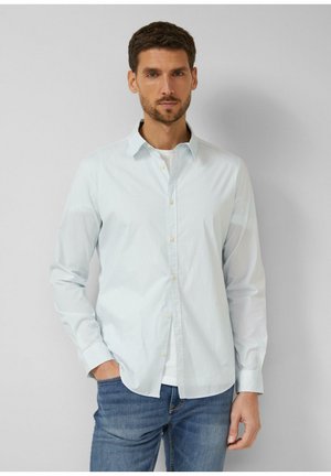 Mężczyzna o krótkich brązowych włosach, ubrany w jasnoniebieską koszulę w paski na guziki, na białym t-shircie i niebieskich dżinsach, stoi na tle gładkiej szarej ściany.