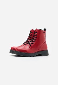 Un par de botas de tobillo lace-up brillantes rojas con suelas negras y tiradores en un fondo blanco.