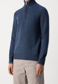 Maglione blu navy con zip al collo e struttura a coste, con dettagli contrastanti azzurri su colletto e polsini. Indossato con pantaloni beige.