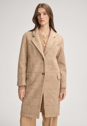 Vrouw draagt een beige geruite wollen jas over een lichtbruine trui met gouden knopen, staand tegen een effen lichte achtergrond.