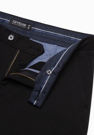 Schwarze Hose mit einem blauen Innenbund. Sie verfügt über einen Reißverschluss, einen Knopfverschluss und einen strukturierten Stoff. Das Etikett trägt die Aufschrift "Ombre Quintessence."
