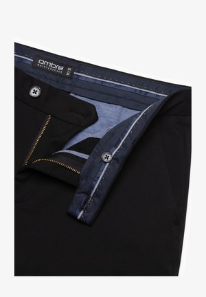 Schwarze Hose mit einem blauen Innenbund. Sie verfügt über einen Reißverschluss, einen Knopfverschluss und einen strukturierten Stoff. Das Etikett trägt die Aufschrift "Ombre Quintessence."