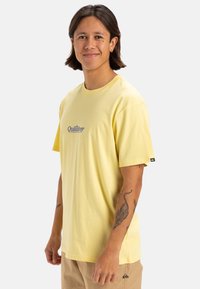 Camiseta de algodón amarilla de manga corta, con un logo de Quiksilver en azul en el pecho y una etiqueta negra en la manga izquierda. Corte regular.