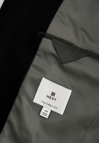 Doublure intérieure de la veste avec étiquette blanche cousue indiquant « NEXT Tailored Fit » et étiquette de taille « 40R Made in Vietnam » sur un tissu gris lisse.
