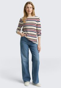 Vrouw staat met hand in broekzak, draagt een gestreepte longsleeve, blauwe wijde jeans en witte sneakers op een effen achtergrond.